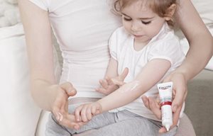 Dievčatko sedí mamičke v lone a natiera si Eucerin AtopiControl Acute krém na ohyb ruky.